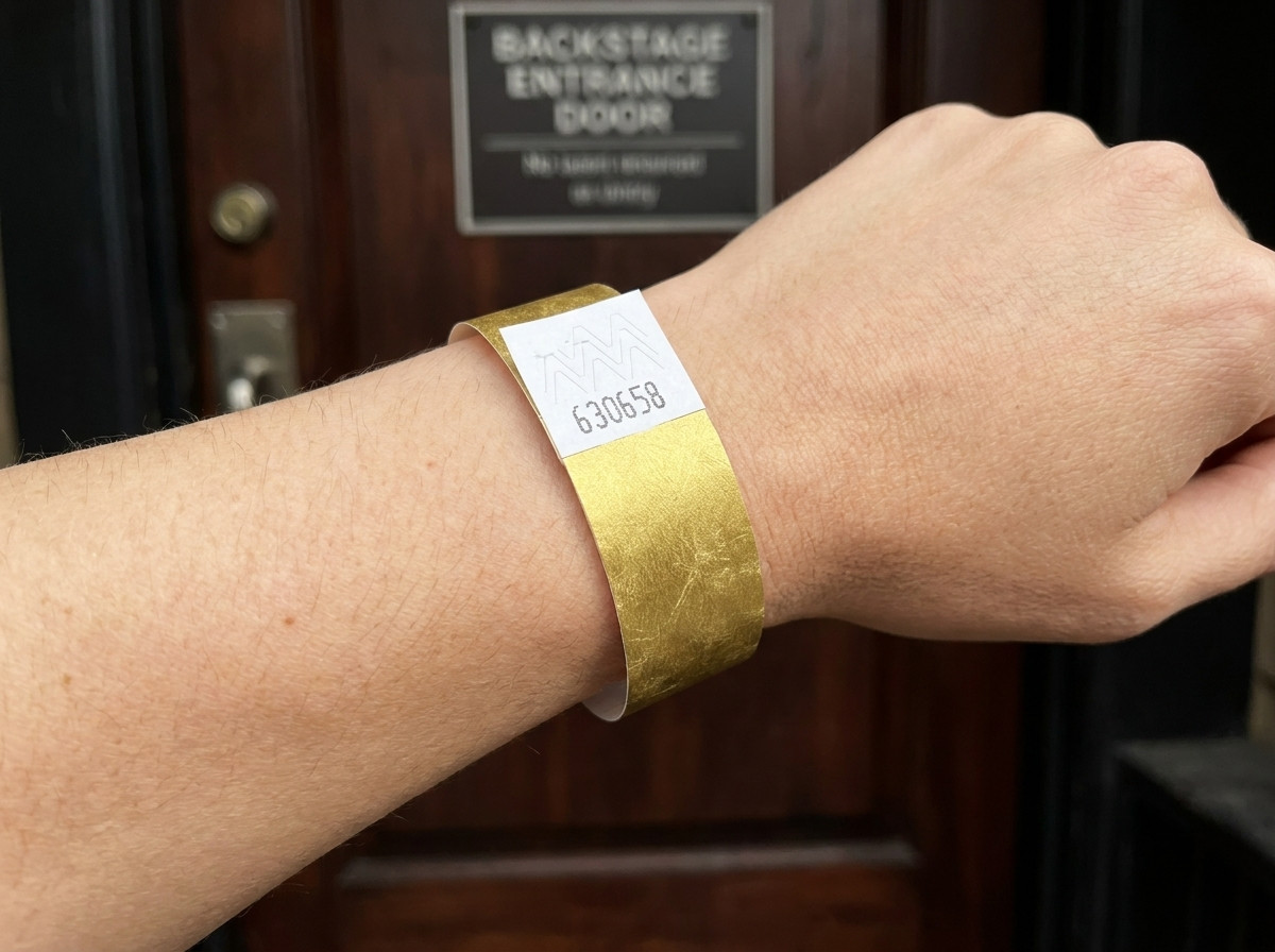 VIP-festivalarmbånd med guldmetalliktryk og stregkode, båret på et håndled foran en backstage-indgang